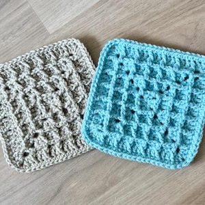 Puede incluir: Dos paños de crochet cuadrados. Uno es gris claro, el otro azul claro. Ambos tienen un patrón texturizado y tejido. Los paños están hechos a mano y se encuentran sobre una superficie de madera clara.