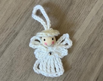 Op de afbeelding: Een handgemaakte witte gehaakte engelversiering met een houten kralen hoofd, roze wangen en een kleine edelsteen. De engel heeft een halo en vleugels, en een lus om op te hangen. De versiering ligt op een houten oppervlak.