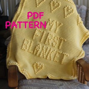 Op de afbeelding: Een gele gehaakte babydeken met de woorden "Happy First Anniversary" in reliëf. De deken heeft een gegolfde rand en ligt op een houten stoel. De tekst "PDF PATTERN" is over de afbeelding gelegd.