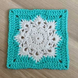 May include: A square, handmade crochet piece featuring a white floral design in the center, surrounded by a turquoise border. The yarn appears soft and the stitches are tight, creating a textured surface. The piece is likely a decorative item or part of a larger craft project.