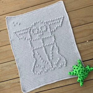Pode incluir: Um cobertor de bebé em crochet cinzento com um design texturizado de uma nave espacial. O cobertor está sobre uma superfície de madeira com um brinquedo de sapo verde no lado direito.