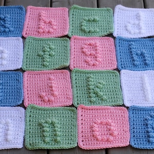 May include: A collection of 12 crocheted squares in various colors, including blue, pink, green, and white. Each square features a different letter of the alphabet, creating a unique and colorful alphabet set.