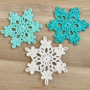 May include: Three handmade crochet snowflakes in shades of teal and white. The snowflakes have a delicate, openwork design with six points. The snowflakes are arranged on a light brown wooden surface.