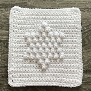 Puede incluir: Cuadrado de crochet blanco con un patrón de diamante en relieve en el centro.