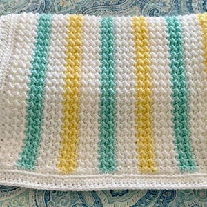 Op de afbeelding: Een handgemaakte gehaakte deken met verticale strepen in wit, lichtblauw en geel. De deken heeft een gestructureerd steekpatroon en een witte rand. Deze zachte deken is ideaal voor een baby of als decoratief kleed.