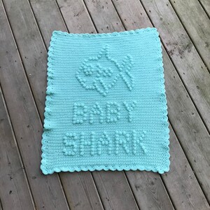 May include: A light blue crocheted baby blanket with a white crocheted border. The blanket has a raised design of leaves and the word "remarkable" in the center.