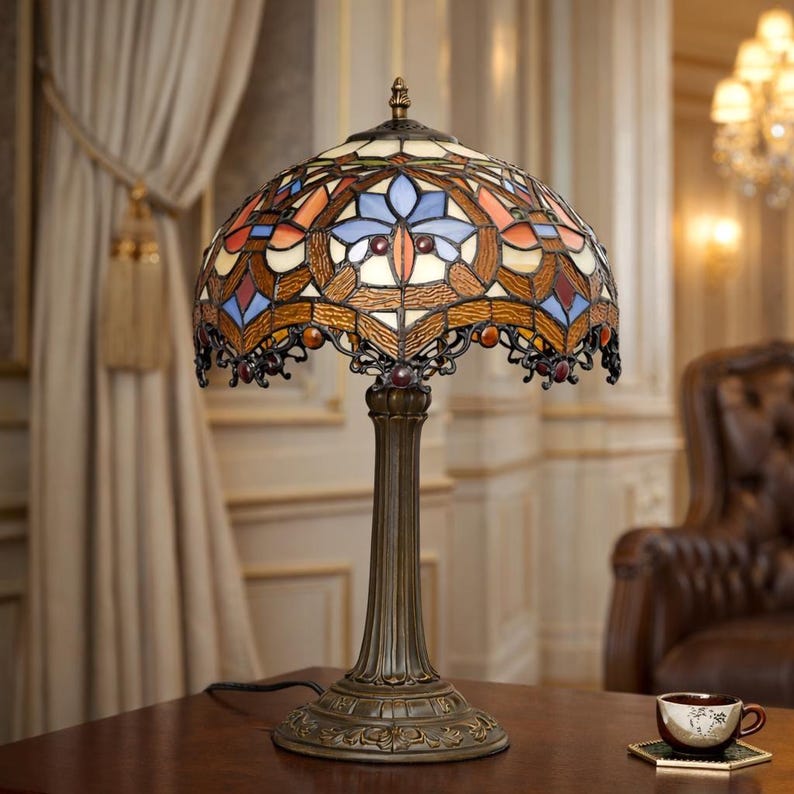 K&ouml;nnte beinhalten: Eine Tischlampe im Tiffany-Stil mit einem Buntglasschirm mit floralem Design in Blau-, Orange- und Wei&szlig;t&ouml;nen. Die Lampe hat einen bronzefarbenen Sockel und ein verziertes Design. Die Lampe steht auf einem Holztisch.