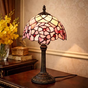May include: A stained glass table lamp with a floral design. The lamp features a pink and white shade with red accents, supported by a dark bronze base and stem. The lamp is on a wooden surface.
