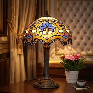 May include: A stained glass table lamp with a domed shade featuring a colorful geometric design in shades of blue, yellow, orange, and red. The lamp has a bronze-colored base and stem. A small white flower pot with pink flowers sits nearby.