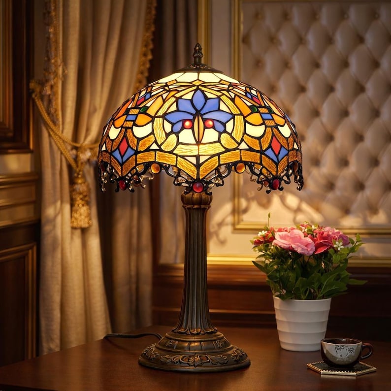 K&ouml;nnte beinhalten: Eine Tischlampe aus Buntglas mit einem gew&ouml;lbten Schirm mit einem farbenfrohen geometrischen Design in Blau-, Gelb-, Orange- und Rott&ouml;nen. Die Lampe hat einen Sockel und einen Stiel in Bronzefarbe. Ein kleiner wei&szlig;er Blumentopf mit rosa Blumen steht daneben.