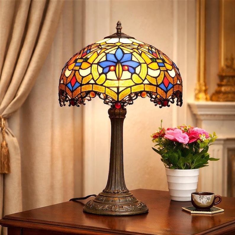 K&ouml;nnte beinhalten: Eine Tischlampe im Tiffany-Stil mit einem Buntglasschirm in Gelb-, Blau-, Rot- und Oranget&ouml;nen. Die Lampe hat einen Sockel in Bronzefarbe und einen dekorativen, gewellten Rand. Ein kleiner Strau&szlig; rosa Blumen steht daneben.