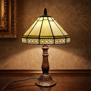 May include: A stained glass table lamp with a geometric design. The lamp features a yellow and cream-colored shade with black outlines and a decorative border. The base is a dark brown color with ornate detailing.