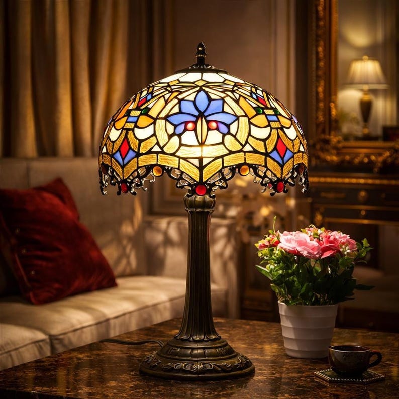 K&ouml;nnte beinhalten: Eine Tiffany-Tischlampe mit einem Sockel aus Bronze und einem farbenfrohen, gew&ouml;lbten Schirm. Der Schirm zeigt ein florales Design mit gelben, blauen, roten und gr&uuml;nen Glasteilen. Die Lampe steht auf einem marmor&auml;hnlichen Tisch, mit einem Sofa und Blumen im Hintergrund.