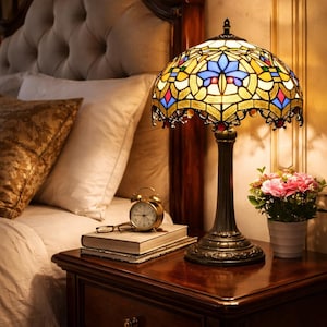 May include: A stained glass table lamp with a bronze base and a colorful shade sits on a wooden nightstand. The lamp's shade features blue, yellow, and red glass pieces. A small alarm clock, glasses, and a book are also on the nightstand.