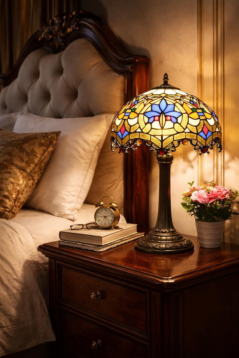 K&ouml;nnte beinhalten: Eine Buntglas-Tischlampe mit einem Sockel aus Bronze und einem farbenfrohen Schirm. Die Lampe steht auf einem Nachttisch aus Holz, neben einem kleinen Wecker und einem Buch. Der Lampenschirm zeigt ein Mosaik aus blauem, gelbem und rotem Glas.
