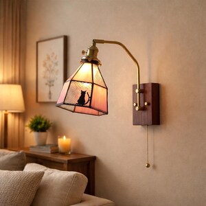 May include: A wall-mounted lamp with a stained glass shade featuring a black cat silhouette. The lamp has a brass arm and a wooden base. The shade is pink and the lamp is turned on, illuminating the room.