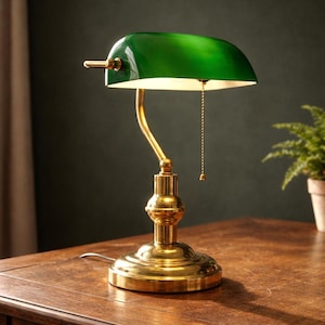 May include: A classic banker's lamp with a green glass shade and a brass base. The lamp features a curved arm and a pull chain for easy operation. It sits on a wooden surface, providing warm, ambient lighting.