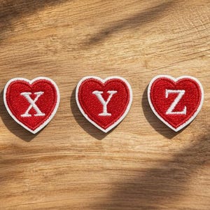 May include: Three red heart-shaped patches with white embroidered borders and letters X, Y, and Z. The patches are on a wooden surface, with a candle and dried flowers in the background.