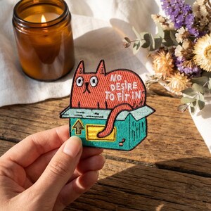 May include: A red cat patch with the text "NO DESIRE TO FIT IN" is shown. The cat is in a teal box with an arrow. A lit candle in a brown jar and dried flowers are in the background.