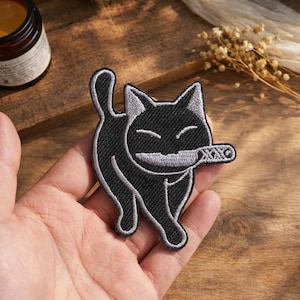 Può includere: Toppa ricamata a forma di gatto nero con dettagli grigi, raffigurante un gatto sorridente che tiene un piccolo oggetto in bocca. La toppa ha un design giocoso, adatta per abbigliamento o accessori.