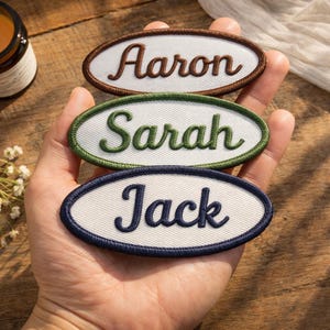 May include: Three embroidered name patches in an oval shape. The names "Aaron", "Sarah", and "Jack" are stitched in a cursive font on a white background. Each patch has a different colored border: brown, green, and navy blue.