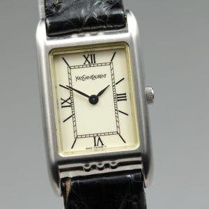 Puede incluir: Un reloj de pulsera rectangular con caja de metal plateado y correa de cuero negro texturizado. La esfera de color blanco roto presenta números romanos negros, manecillas negras y el texto Yves Saint Laurent con un número de serie.