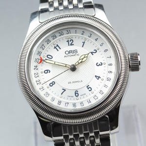 Może przedstawiać: Srebrny automatyczny zegarek na rękę ORIS z białą tarczą, czarnymi cyframi i wskazówkami. Zegarek posiada wyświetlacz daty i teksturowaną ramkę. Pasek wykonany jest z połączonego metalu. Na tarczy zegarka widnieje napis "ORIS AUTOMATIC 25 JEWELS".