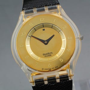Puede incluir: Un reloj Swatch dorado con una correa negra. La esfera del reloj tiene números y agujas doradas, con el logotipo de Swatch en el centro. La caja del reloj es de plástico transparente. El reloj está marcado "SWATCH SWISS" y "© SWATCH AG 2002".