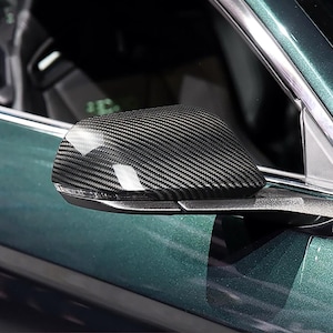 May include: A carbon fiber side mirror on a green car. The mirror has a glossy finish and a woven pattern. The car's body is a dark green color, and the window frame is silver.