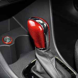 Op de afbeelding: Een rode koolstofvezel auto versnellingspookknop met een chromen accent. De versnellingspook bevindt zich in een zwart interieur. De knop heeft een glanzende afwerking en een gestructureerd patroon. De startknop van de auto is zichtbaar op de achtergrond.