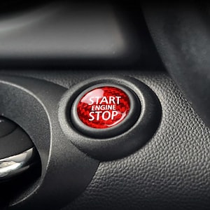 May include: A close-up of a car dashboard showing a red "START ENGINE STOP" button. The button has a textured, carbon fibre-like appearance and is set in a black plastic panel. The circular button is clearly visible.