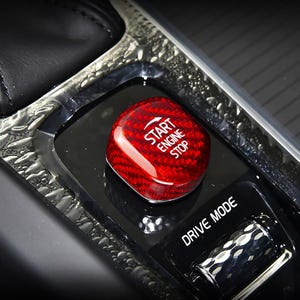 May include: A red, carbon fiber-textured start/stop button with white text. The button is in a black console with the words "DRIVE MODE" visible. The button is rectangular with rounded edges.