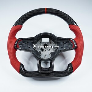 Puede incluir: Un volante de fibra de carbono con empuñaduras de cuero perforado rojo. La parte superior del volante tiene una franja roja. El centro del volante tiene una carcasa de plástico negro. El identificador de Instagram @xiaoming_jim es visible en el centro del volante.