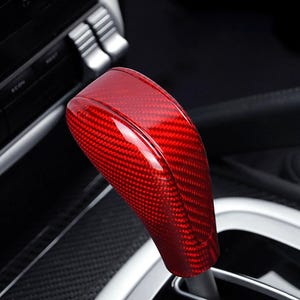 Könnte beinhalten: Ein roter Schalthebelknauf aus Kohlefaser. Der Schalthebelknauf hat eine glänzende Oberfläche und ein gewebtes Muster. Der Hintergrund ist der Innenraum eines Autos mit schwarzen und silbernen Akzenten.