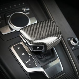 Könnte beinhalten: Nahaufnahme eines Autoschalthebels. Der Schalthebelknauf ist mit einem Karbonfasermuster bedeckt. Der Gangwählhebel zeigt die Buchstaben "R", "N", "D/S" und den Buchstaben "P" für Parken.