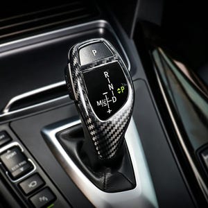 May include: Close-up of a car's gear shift lever with a carbon fiber pattern. The lever displays the gear positions: P, R, N, D, and M/S. The background shows the car's interior with buttons and a silver trim.