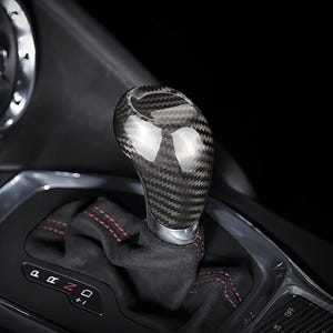 May include: A carbon fiber gear shift knob with a glossy finish. The shift knob is installed in a vehicle's interior, with a black and red stitched shift boot visible. The gear selector shows the positions P, R, N, D, and +1.