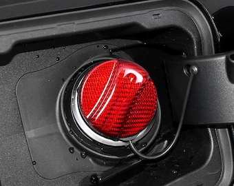 May include: A circular red fuel cap with a textured carbon fiber pattern, featuring a glossy finish and a black tether cord. The cap is seated within a black plastic fuel filler housing, which is integrated into the exterior of a vehicle body.