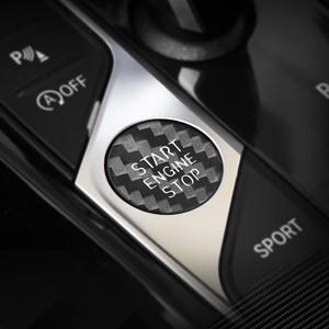 May include: A circular engine start stop button featuring a black and gray carbon fiber pattern. The button is set within a silver metallic trim on a vehicle console, with white text reading START ENGINE STOP centered on the button surface.
