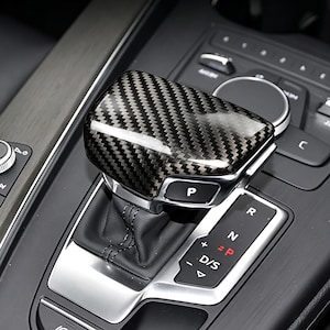 May include: A carbon fiber automatic gear shift knob with a silver base. The knob is in the "P" (Park) position. The surrounding console is black with silver accents and other controls.
