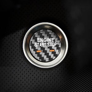 May include: A circular car engine start/stop button with a silver metallic rim. The button's center features a black and gray carbon fiber pattern with the words "ENGINE START/STOP" in white. Two orange horizontal lines are below the text.