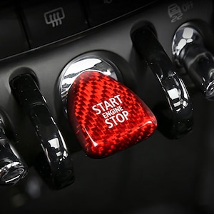 May include: A close-up of a car's dashboard featuring a red, textured start/stop button with the words "START ENGINE STOP" in white. The button is surrounded by black levers and other controls, with a focus on the button's design.
