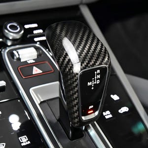 Könnte beinhalten: Ein Schalthebel aus Kohlefaser mit strukturiertem Muster. Der Hebel hat ein schwarz-graues Design mit weißer Beschriftung, die die Gangpositionen angibt. Der Hebel befindet sich in der Position 'P' (Parken), mit einer roten Kontrollleuchte.