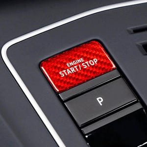 May include: Close-up of a car's dashboard featuring a red carbon fiber-textured engine start/stop button with white lettering. Below it is a black button with a white "P" for park. The surrounding panel is dark gray.