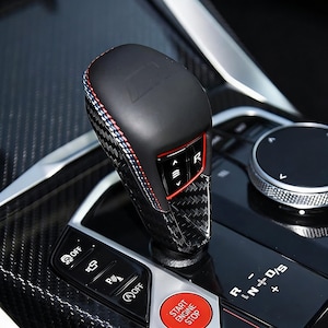 Può includere: Pomello del cambio auto nero e in fibra di carbonio con cuciture rosse, bianche e blu. La manopola ha un accento rosso e la lettera "M" in alto. Il cruscotto dell'auto include un pulsante start/stop.