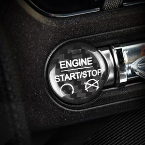 May include: A black and silver engine start/stop button with a carbon fiber pattern. The button has white text that reads "ENGINE START/STOP" and a power symbol. The button is part of a car's dashboard.