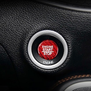 May include: A close-up of a car's engine start/stop button. The button is red with a carbon fiber pattern and white text that reads "ENGINE START STOP" and "ENGINE". The button is surrounded by a silver ring and set in a black leather interior.