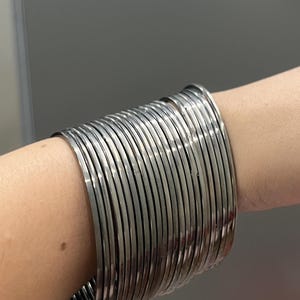 May include: A set of numerous thin, silver-toned metal bangles worn together on the wrist. The bracelets are smooth, circular, and have a reflective, polished finish, creating a wide, stacked appearance across the forearm.