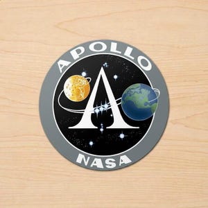 May include: Round gray sticker featuring the Apollo NASA logo. The design includes a stylized letter "A" with a crescent moon and Earth on either side, set against a starry background. The words "APOLLO" and "NASA" are printed around the edge.