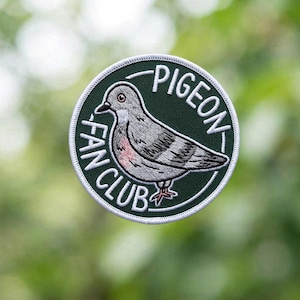 May include: A round, green patch with the words "PIGEON FAN CLUB" in white lettering. The patch features a detailed illustration of a gray pigeon with pink accents, outlined in white.